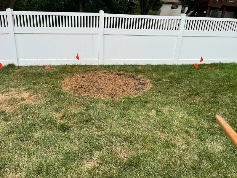 Mound of fresh wood chips in a green lawn where a stump was removed, white vinyl privacy fence behind, utility flags visible